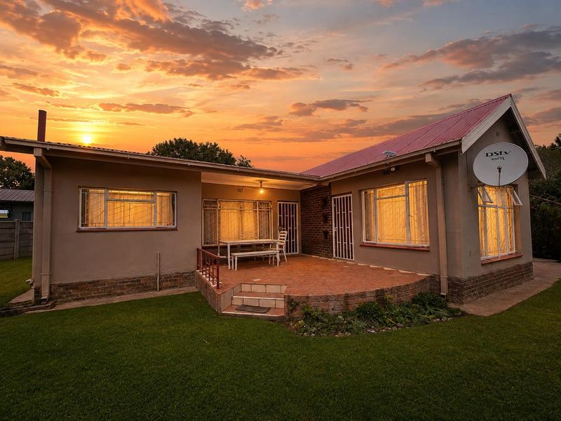 Rear view of the house featuring a brick patio, corrugated metal roof, and security features, set against a sunset sky.