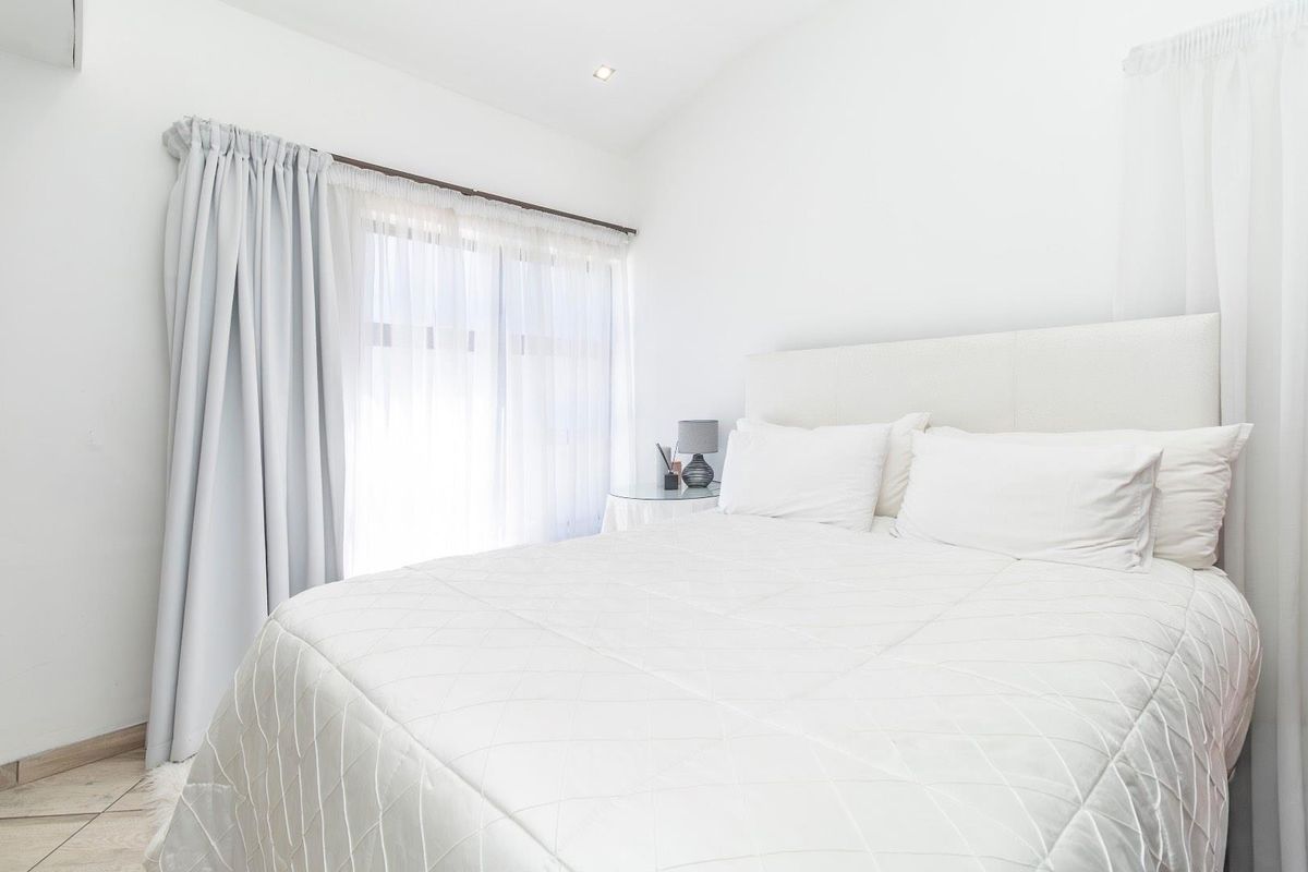 A bright and neutral bedroom featuring tiled flooring, recessed lighting, and large windows dressed with elegant curtains.