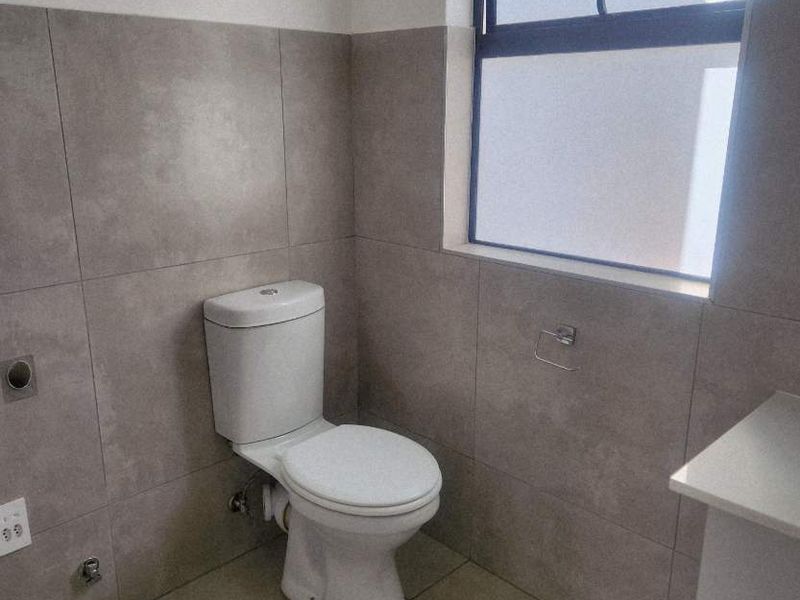 A clean, modern bathroom featuring neutral grey wall and floor tiles, a white toilet, and a functional louvre window for ventilation.
