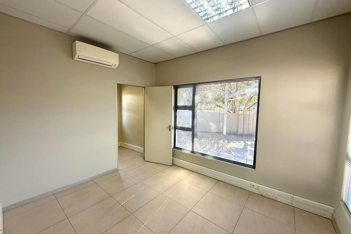 A clean, empty office space featuring tiled flooring, a suspended ceiling with recessed lighting, and a wall-mounted air conditioning unit.