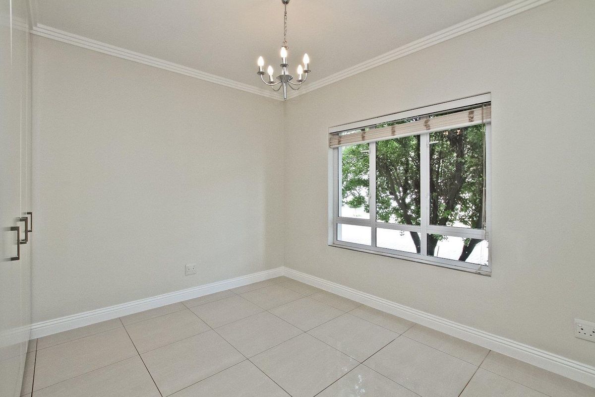 A bright, empty bedroom featuring neutral tiled flooring, built-in cupboards, elegant ceiling cornice, and a central chandelier.