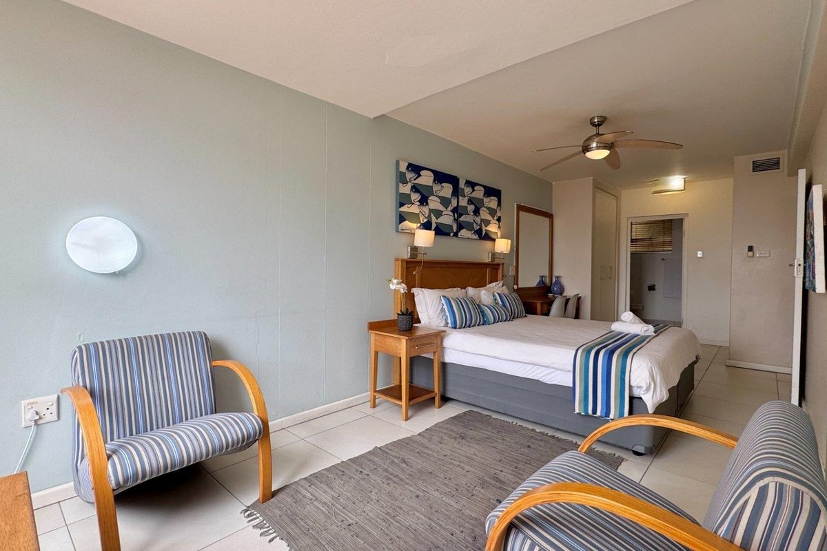 A well-lit bedroom featuring tiled flooring, a ceiling fan, and built-in storage, with an adjoining en-suite bathroom visible in the background.