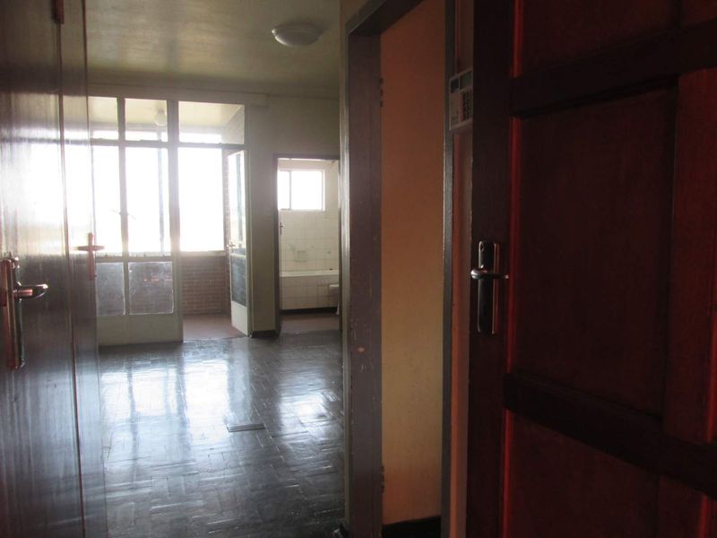 A view through a hallway with parquet flooring, leading towards a sunlit balcony and a glimpse of a bathroom area.
