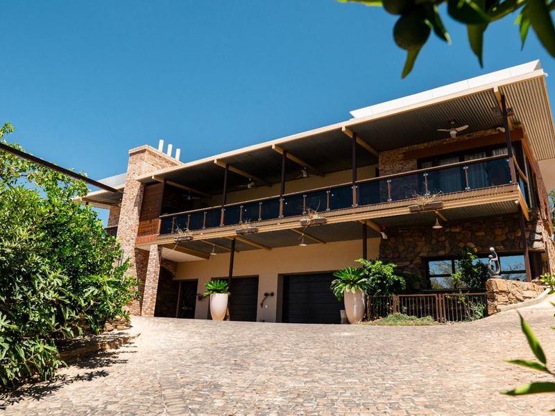 A striking multi-level residence featuring a cobblestone driveway, stone-clad facade, and a spacious covered balcony with glass balustrades.