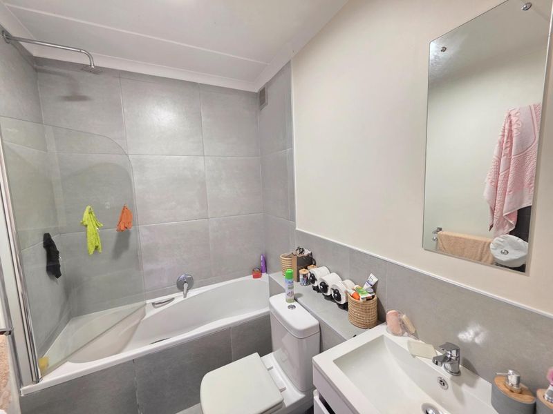 A modern bathroom featuring grey wall tiles, a bathtub with a glass shower screen, a vanity basin, and a wall-mounted mirror.