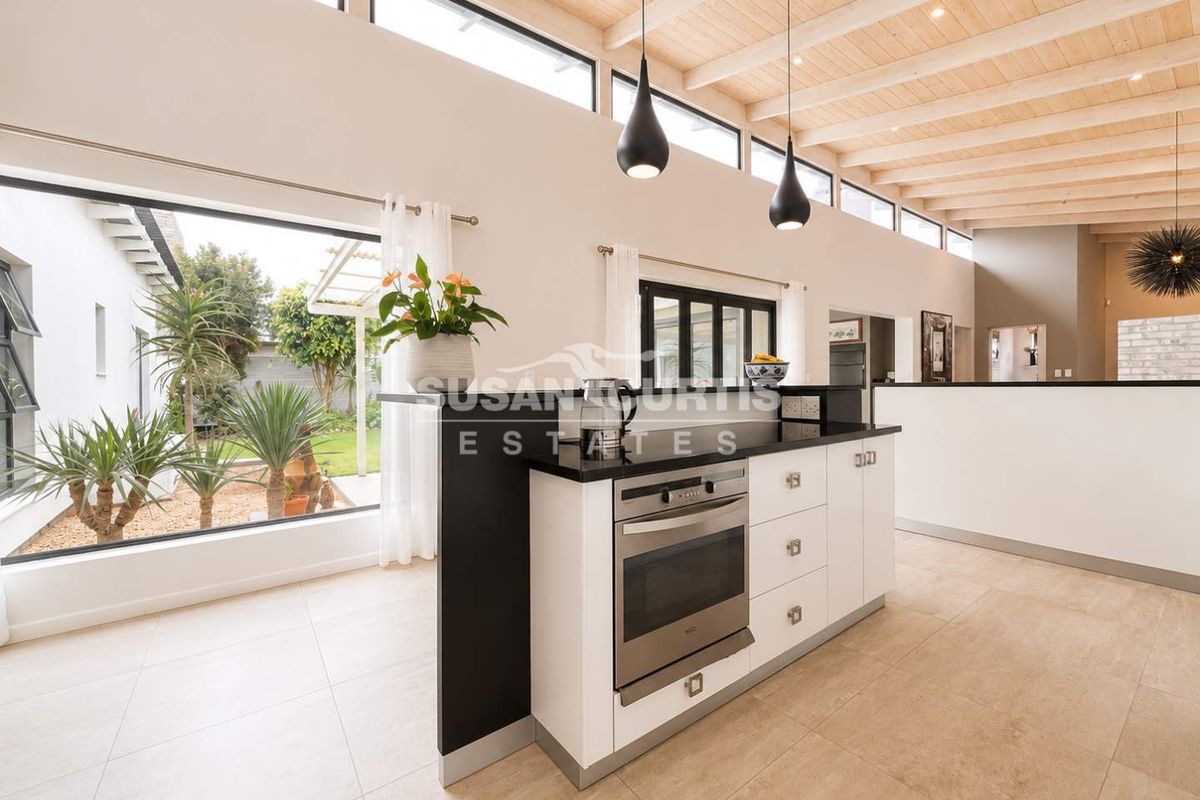 Modern kitchen featuring a central island with built-in oven, sleek black countertops, exposed timber beam ceiling, and large windows overlooking the garden.
