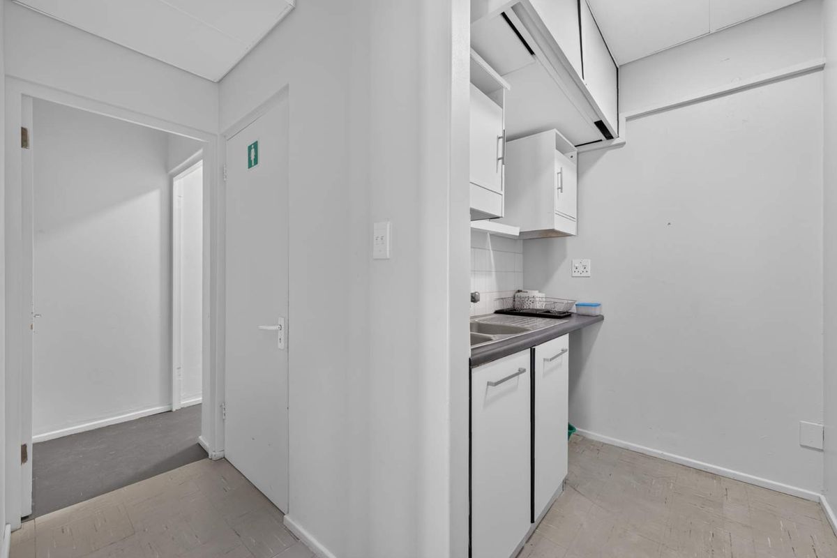 A compact office kitchenette featuring built-in cabinetry, a stainless steel sink, and tiled splashback, adjacent to a hallway with access to multiple rooms.
