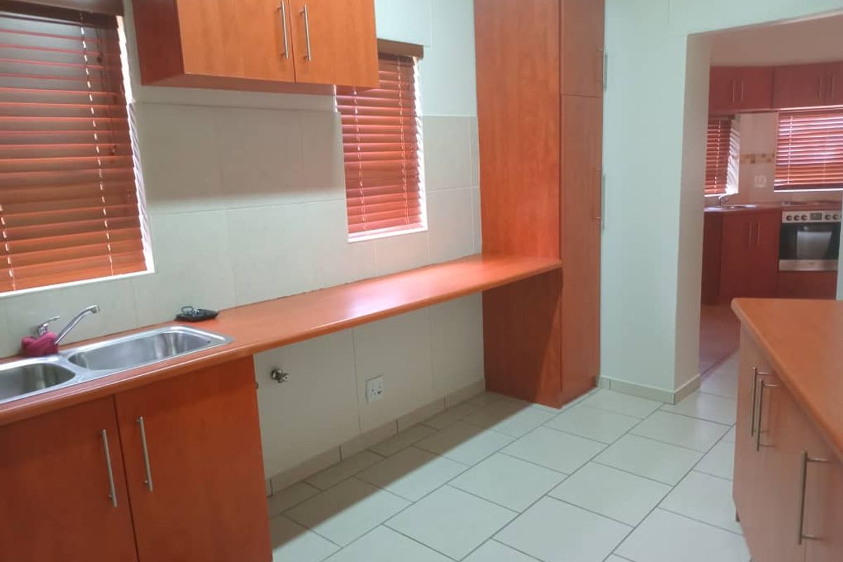 A functional kitchen featuring wood-finish cabinetry, tiled flooring, and a double-basin sink, with an open-plan layout connecting to an adjacent prep area.