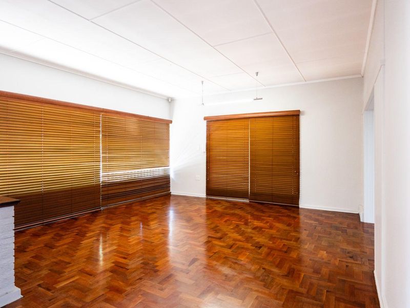 A bright, empty living room featuring classic herringbone parquet flooring and fitted wooden venetian blinds.