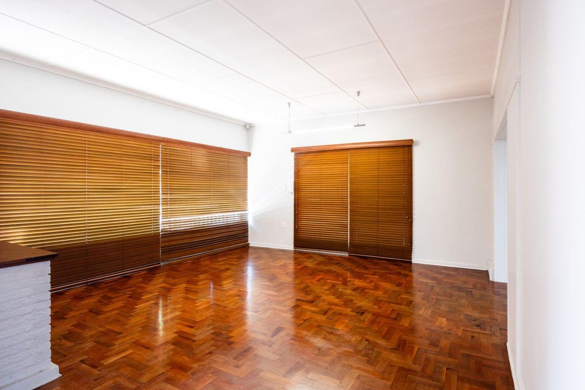 A bright, empty living room featuring classic herringbone parquet flooring and fitted wooden venetian blinds.