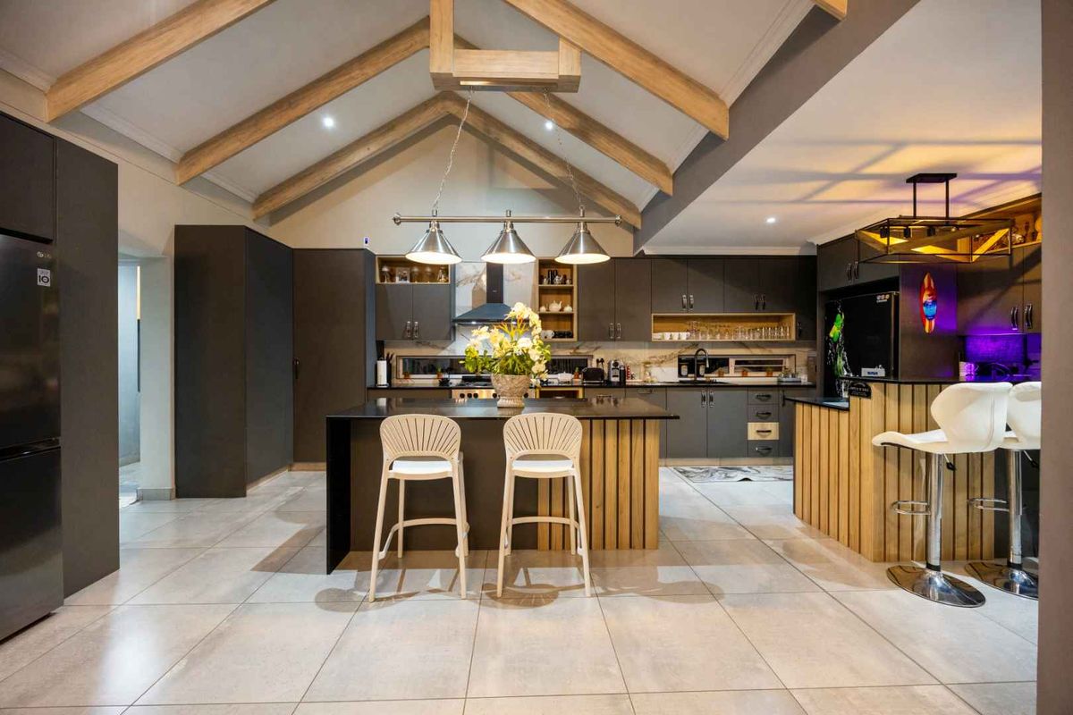 Modern open-plan kitchen featuring a central island, sleek dark cabinetry, vaulted ceilings with exposed timber beams, and high-quality tiled flooring.