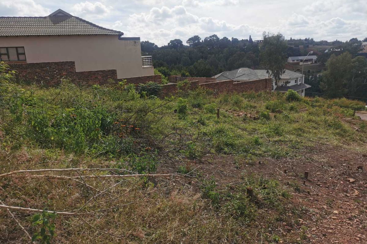 A vacant residential plot featuring natural vegetation and sloping terrain, situated in an established neighborhood with surrounding residential properties.