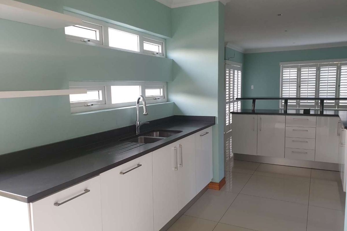 A modern, clean kitchen featuring white cabinetry, dark stone countertops, stainless steel sink, and tiled flooring with plantation shutters.