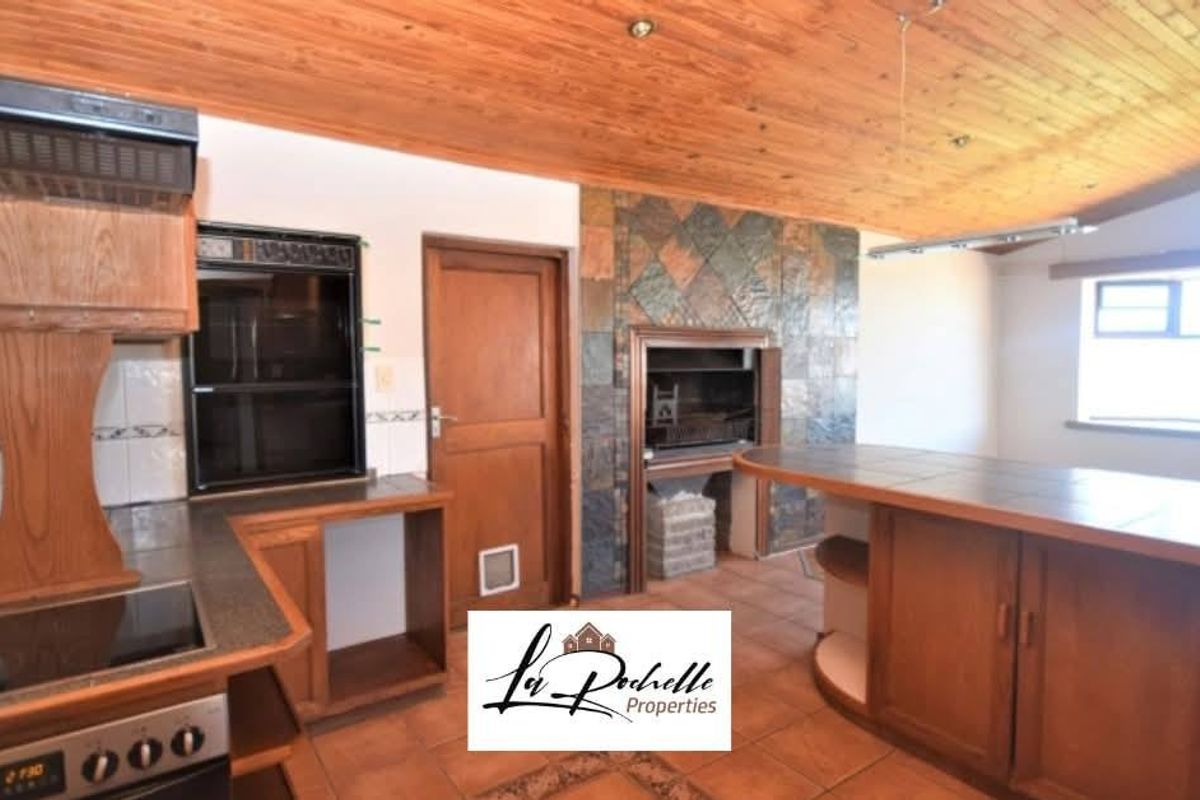 A spacious kitchen featuring wooden cabinetry, a built-in oven, a hob, and a stone-tiled fireplace, with a large breakfast counter and wood-paneled ceiling.