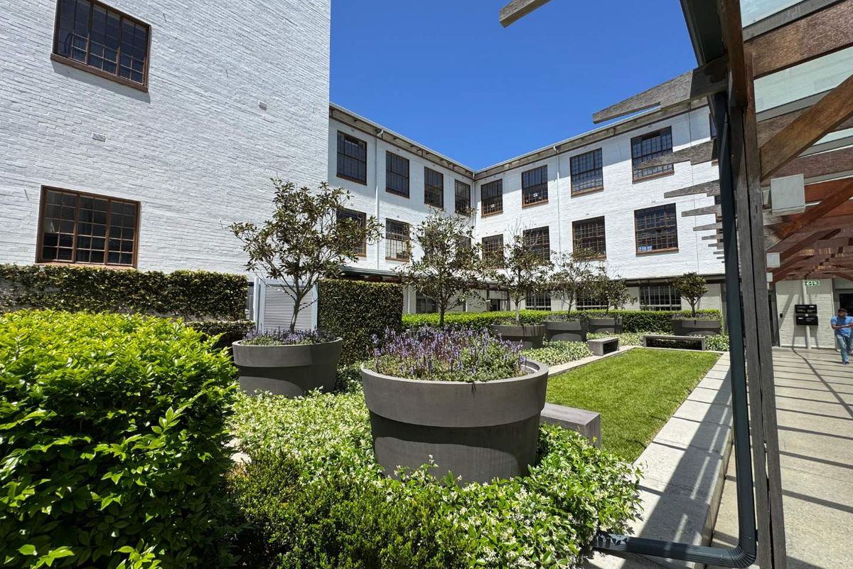 A well-maintained central courtyard featuring landscaped garden beds, manicured lawn, and paved walkways surrounded by a multi-story white brick office building.