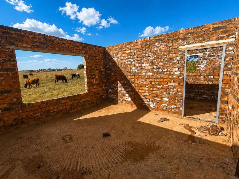 An unfinished brick structure in shell condition, featuring exposed masonry walls and a metal door frame, set within an open rural landscape.