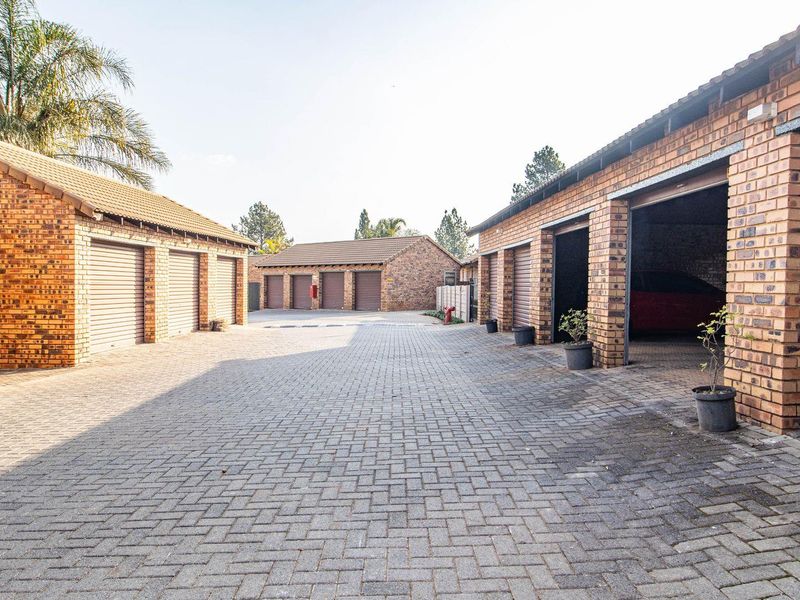 A paved courtyard area featuring multiple brick-built garage units with roller shutter doors in a townhouse complex setting.
