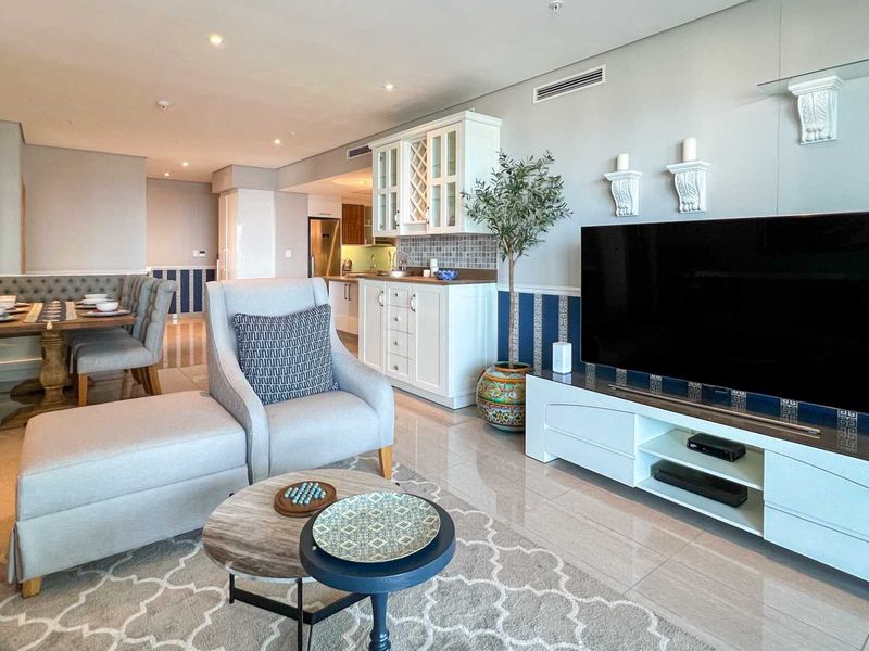 An open-plan living and dining area featuring tiled flooring, recessed lighting, and a built-in kitchenette with white cabinetry and a mosaic backsplash.