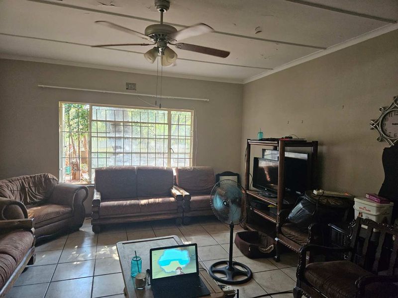 A living room featuring tiled flooring, a ceiling fan, and large windows with security bars. The ceiling shows signs of wear and requires maintenance.