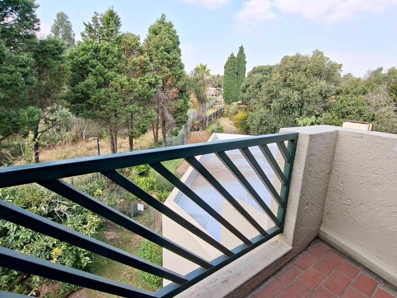 A private balcony featuring a metal balustrade and brick flooring, overlooking a lush garden area with mature trees.