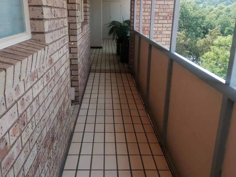 A long, covered balcony featuring exposed brick walls, tiled flooring, and metal railings with views of the surrounding greenery.