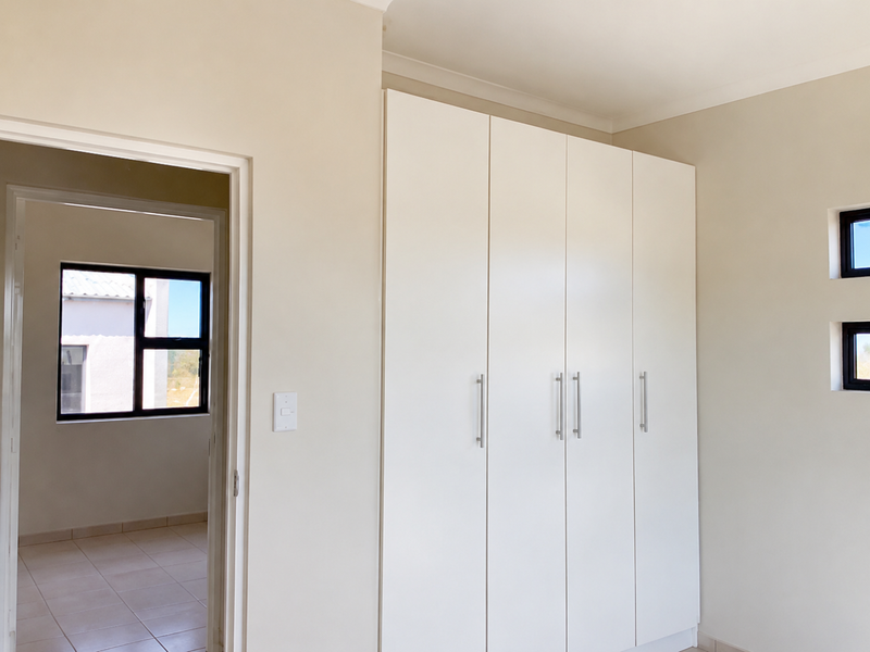 A bright, modern bedroom featuring neutral walls, sleek built-in cupboards, and aluminium-framed windows, offering a clean and versatile living space.