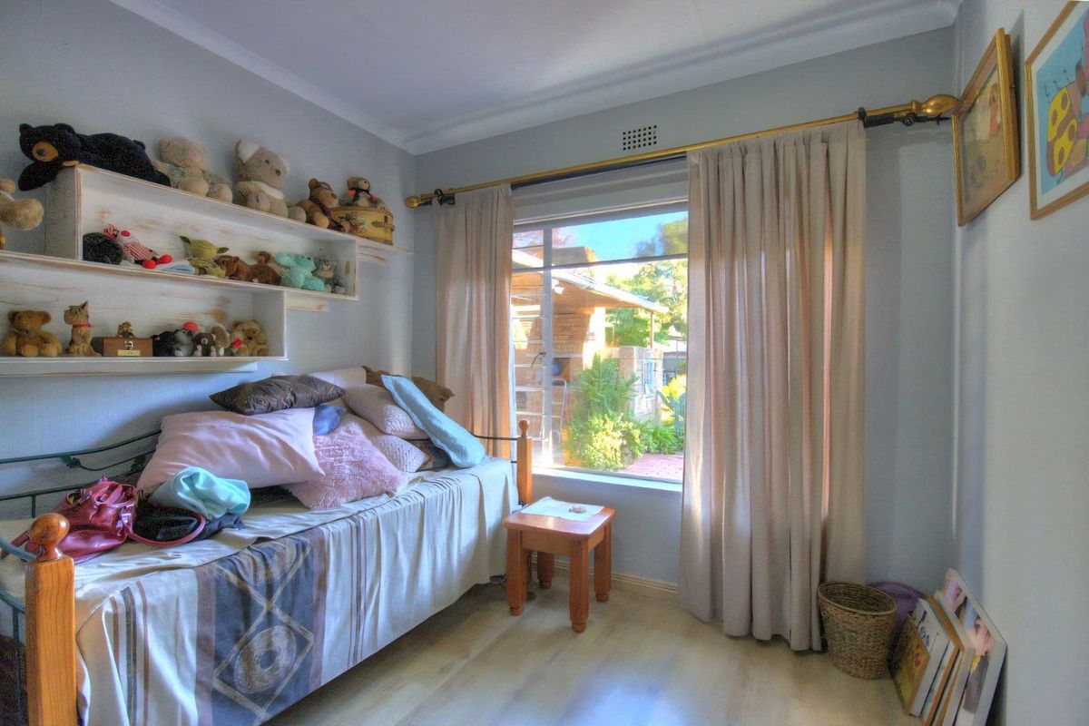 A bedroom featuring light-toned flooring, built-in wall shelving, and a large window with curtain tracks, offering natural light and a view of the garden.