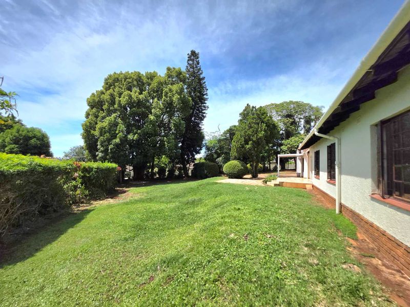 A spacious rear garden featuring a well-maintained lawn, mature trees, and a paved patio area adjacent to the house exterior.
