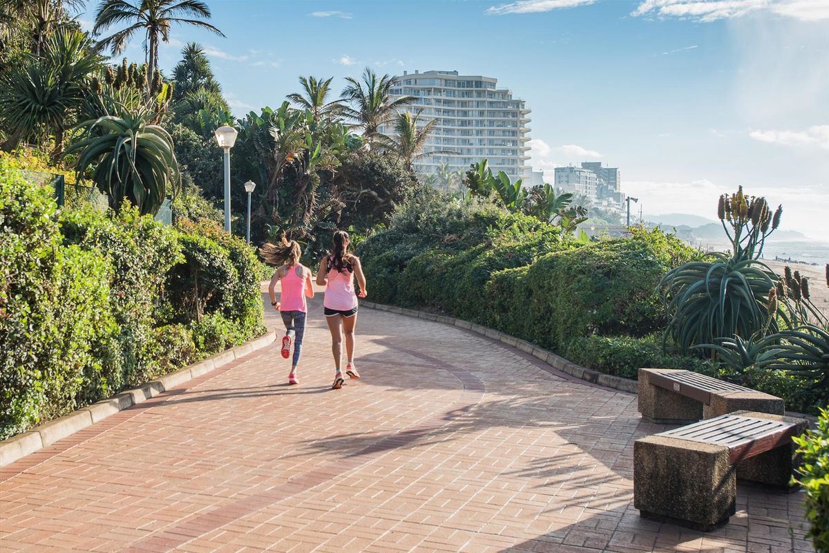 A scenic paved coastal promenade featuring landscaped gardens, street lighting, and built-in stone benches, offering a pleasant outdoor environment near the beach.