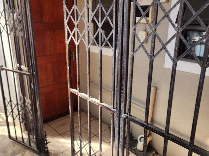 An exterior entrance area featuring a timber door, tiled flooring, and metal security gates showing signs of wear and corrosion.
