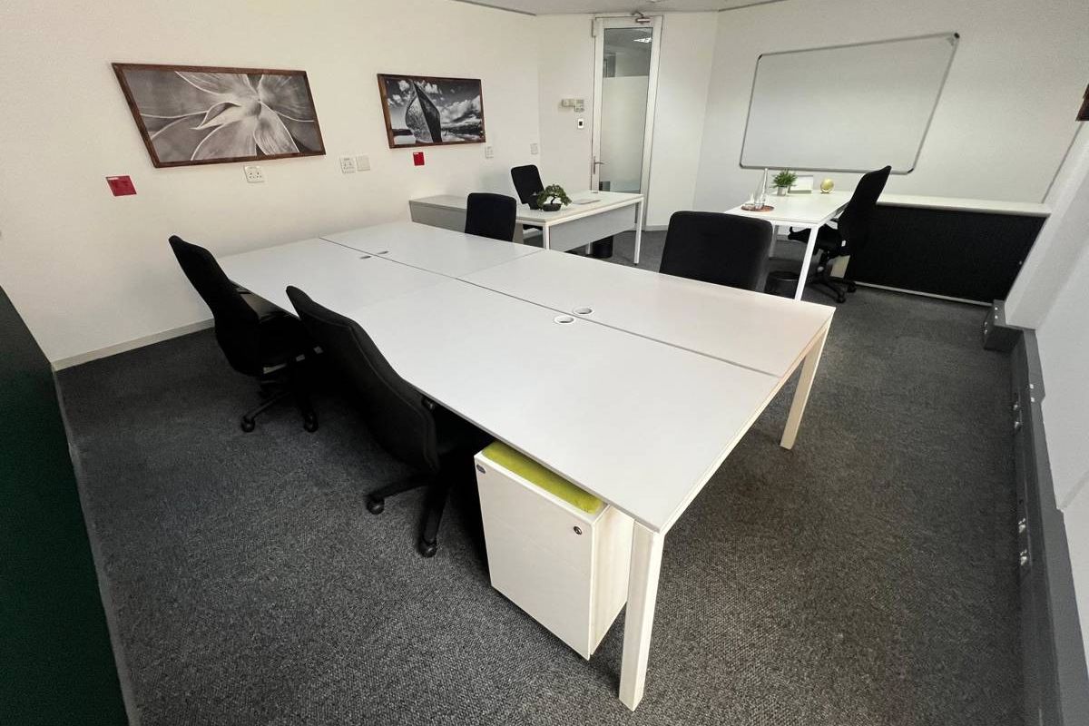 A professional office space featuring carpeted flooring, recessed ceiling lighting, and a wall-mounted whiteboard, providing a functional work environment.