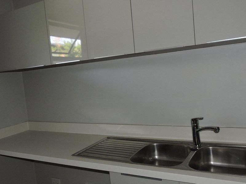 A clean, modern kitchen area featuring white gloss cabinetry, a solid surface countertop, and a stainless steel double sink with a mixer tap.