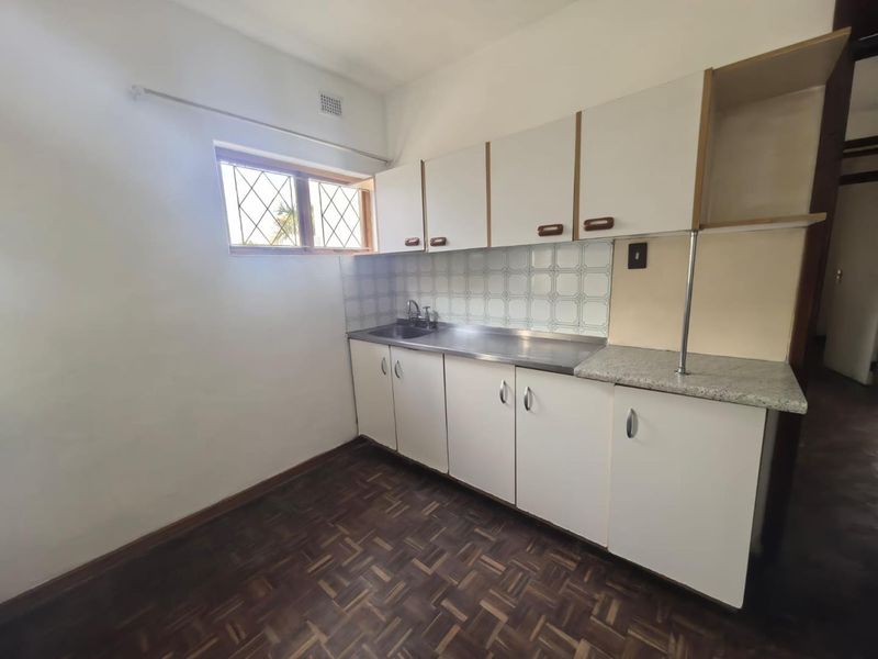 A compact, empty kitchen featuring original parquet flooring, white cabinetry, a stainless steel sink, and a tiled backsplash, offering potential for modern renovation.