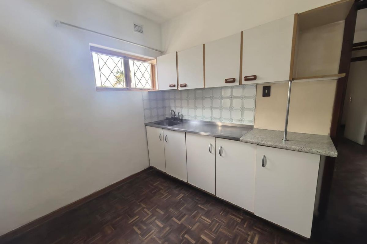 A compact, empty kitchen featuring original parquet flooring, white cabinetry, a stainless steel sink, and a tiled backsplash, offering potential for modern renovation.