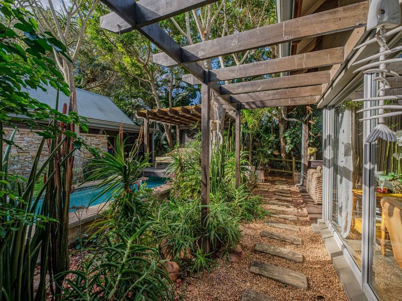 A lush outdoor patio area featuring a timber pergola, stone pathway, and direct access to a swimming pool and garden setting.