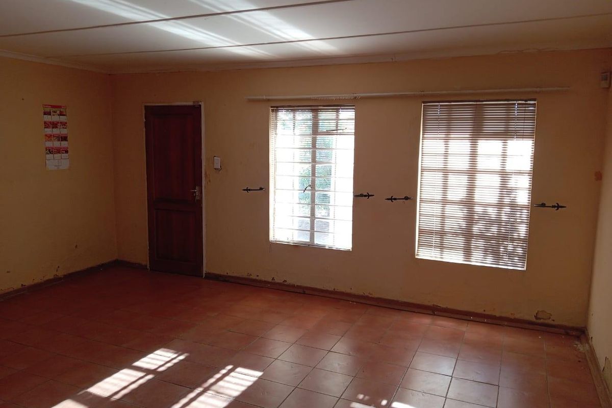 An empty living room featuring tiled flooring, large windows with fitted blinds, and a wooden door, offering a blank canvas for renovation.