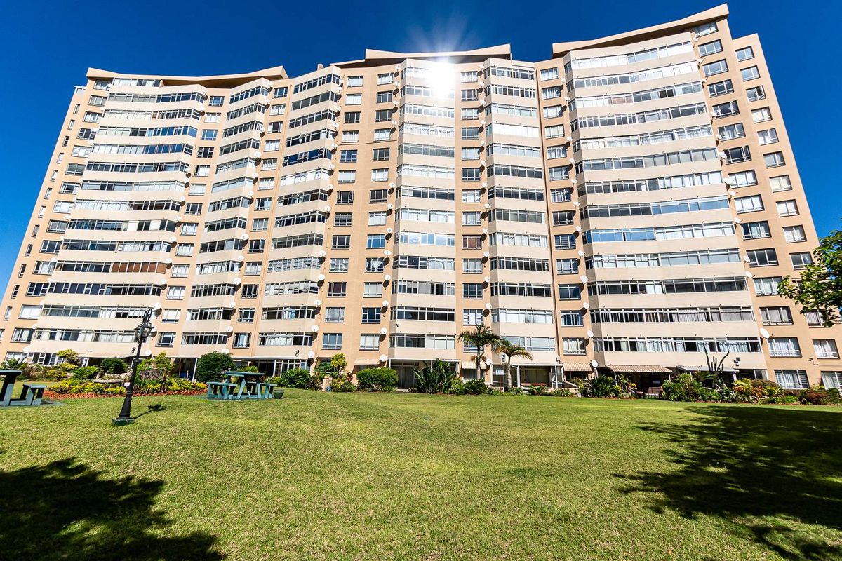 A wide-angle view of the apartment building facade featuring extensive glazing and balconies, set against a well-maintained communal lawn area.