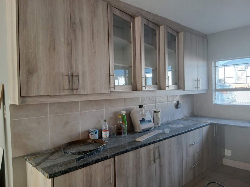 A functional kitchen space featuring light wood-grain cabinetry, glass-fronted upper cupboards, a tiled backsplash, and stone-look countertops, awaiting final finishing touches.