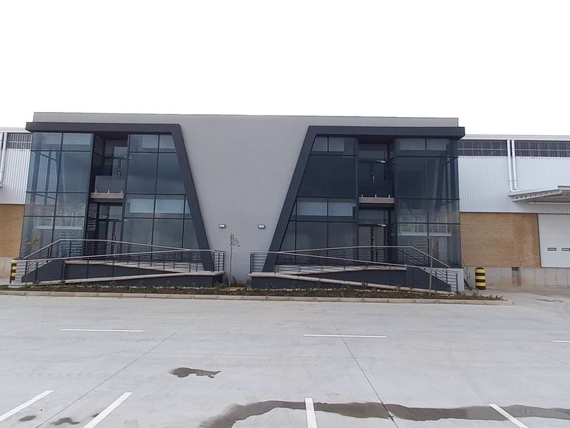 Modern industrial warehouse facility featuring a prominent glass facade, multiple roller shutter loading doors, and a spacious concrete yard area.
