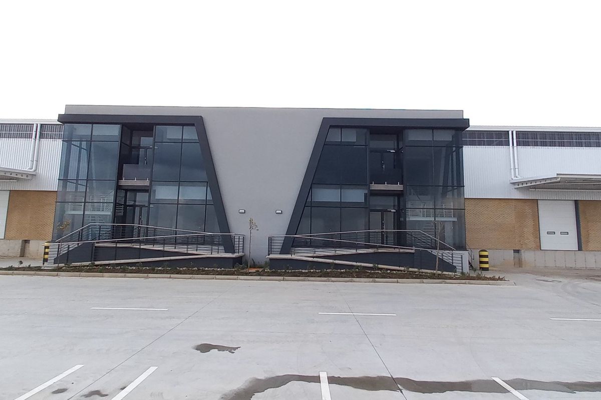 Modern industrial warehouse facility featuring a prominent glass facade, multiple roller shutter loading doors, and a spacious concrete yard area.