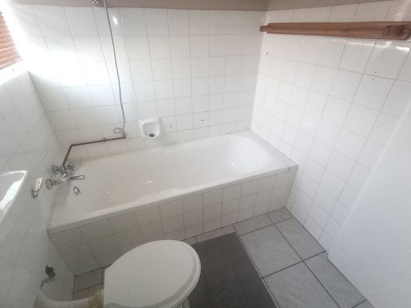A dated bathroom featuring a built-in bathtub with wall-mounted shower mixer, white ceramic wall tiles, and tiled flooring.