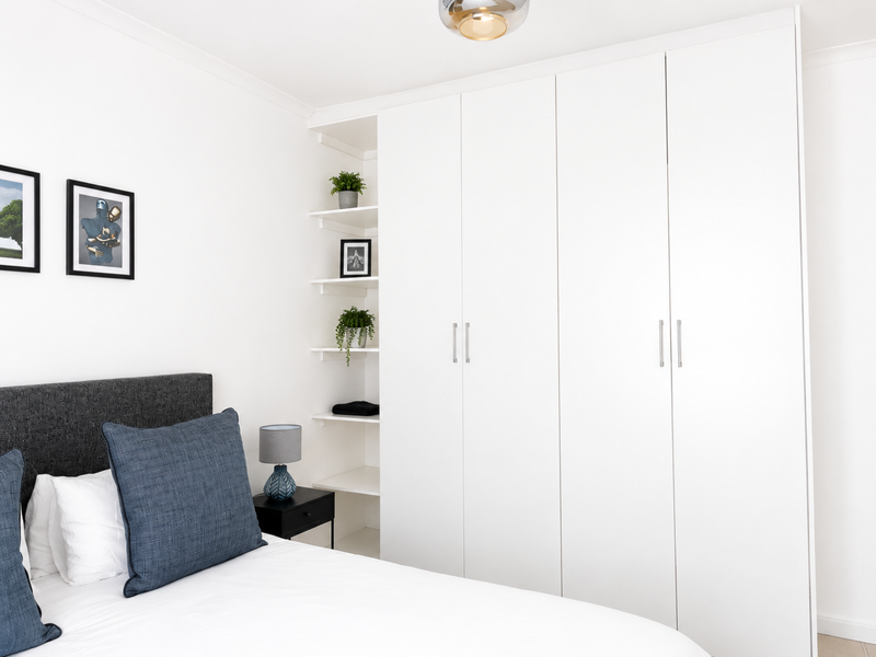 A bright bedroom featuring a large, modern white built-in wardrobe with integrated open shelving and a contemporary ceiling light fixture.