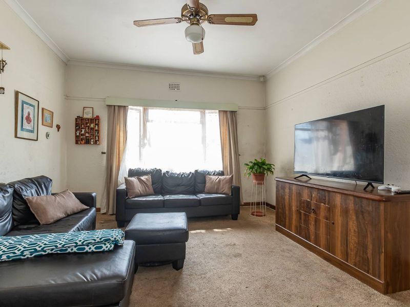 A living room featuring a ceiling fan and a large bay window, currently showing signs of wear on the walls and flooring.