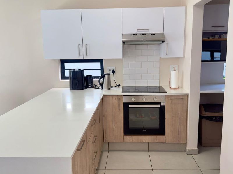 Modern kitchen featuring light wood-finish cabinetry, white stone countertops, subway tile backsplash, and integrated electric cooking appliances with an adjacent scullery area.