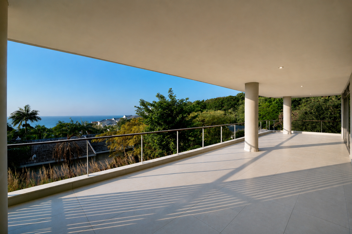 A spacious, modern covered balcony featuring clean tiled flooring, structural columns, and a sleek metal railing with expansive coastal and landscape views.