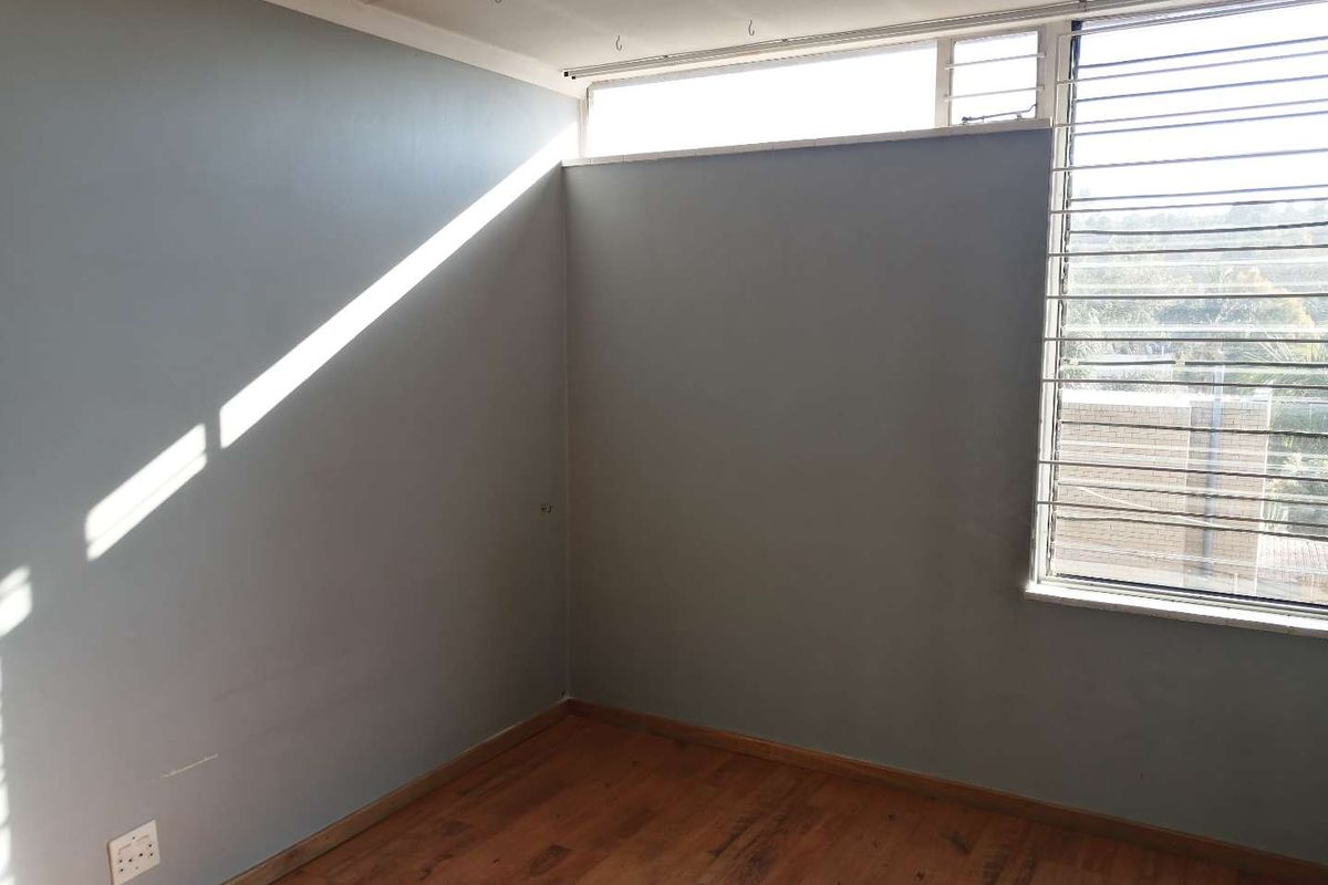 A bright, empty bedroom featuring wooden flooring, large windows with security bars, and a neutral grey wall finish.