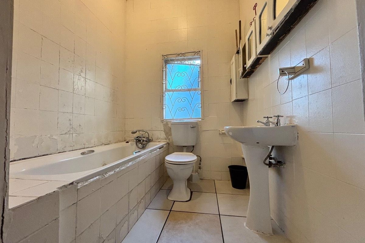 A dated bathroom featuring a bathtub, pedestal sink, and toilet, with significant wear to the wall tiling and finishes throughout.
