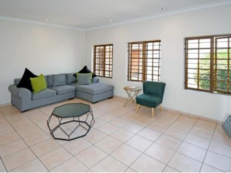 A bright living room featuring neutral tiled flooring, recessed downlighting, and windows fitted with security bars.