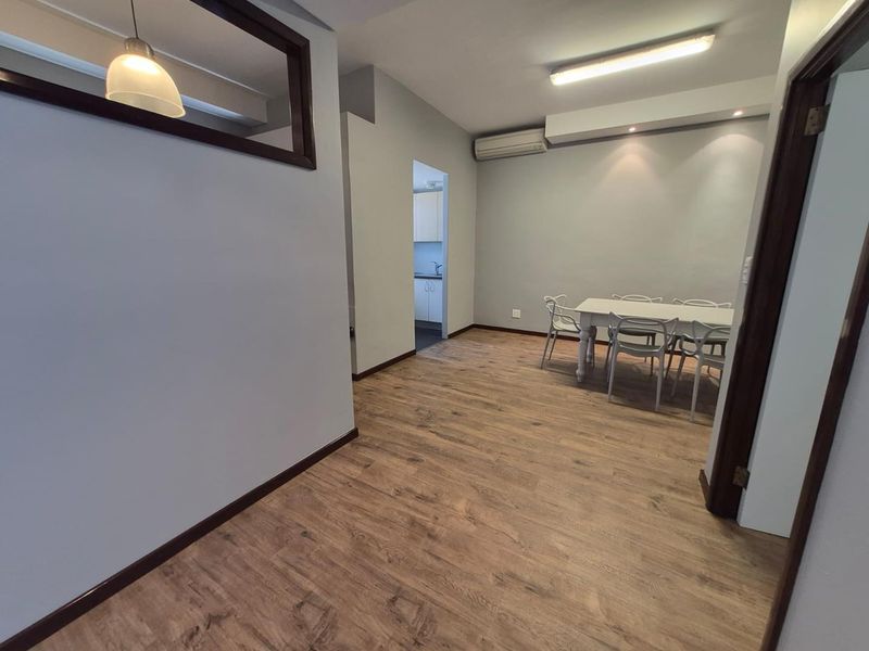 Open-plan office area featuring durable laminate flooring, split-system air conditioning, and a mix of fluorescent and recessed lighting, with an adjacent kitchenette view.