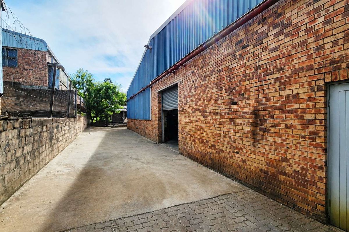 Side exterior view of an industrial property featuring a brick facade, corrugated metal cladding, and a concrete driveway with perimeter security fencing.